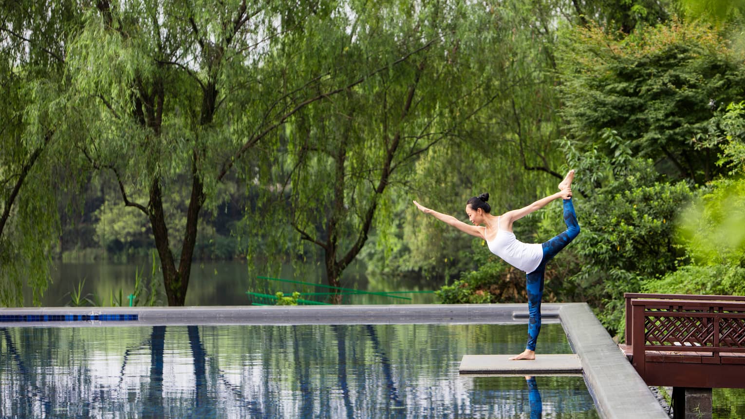 A yogi in a graceful dancer's pose at the edge of a glassy pool, wispy trees reflected in the water, a serene river beyond.