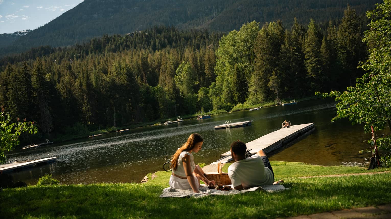 Two people sit on a blanket on the grass having a picnic by a mountain lake