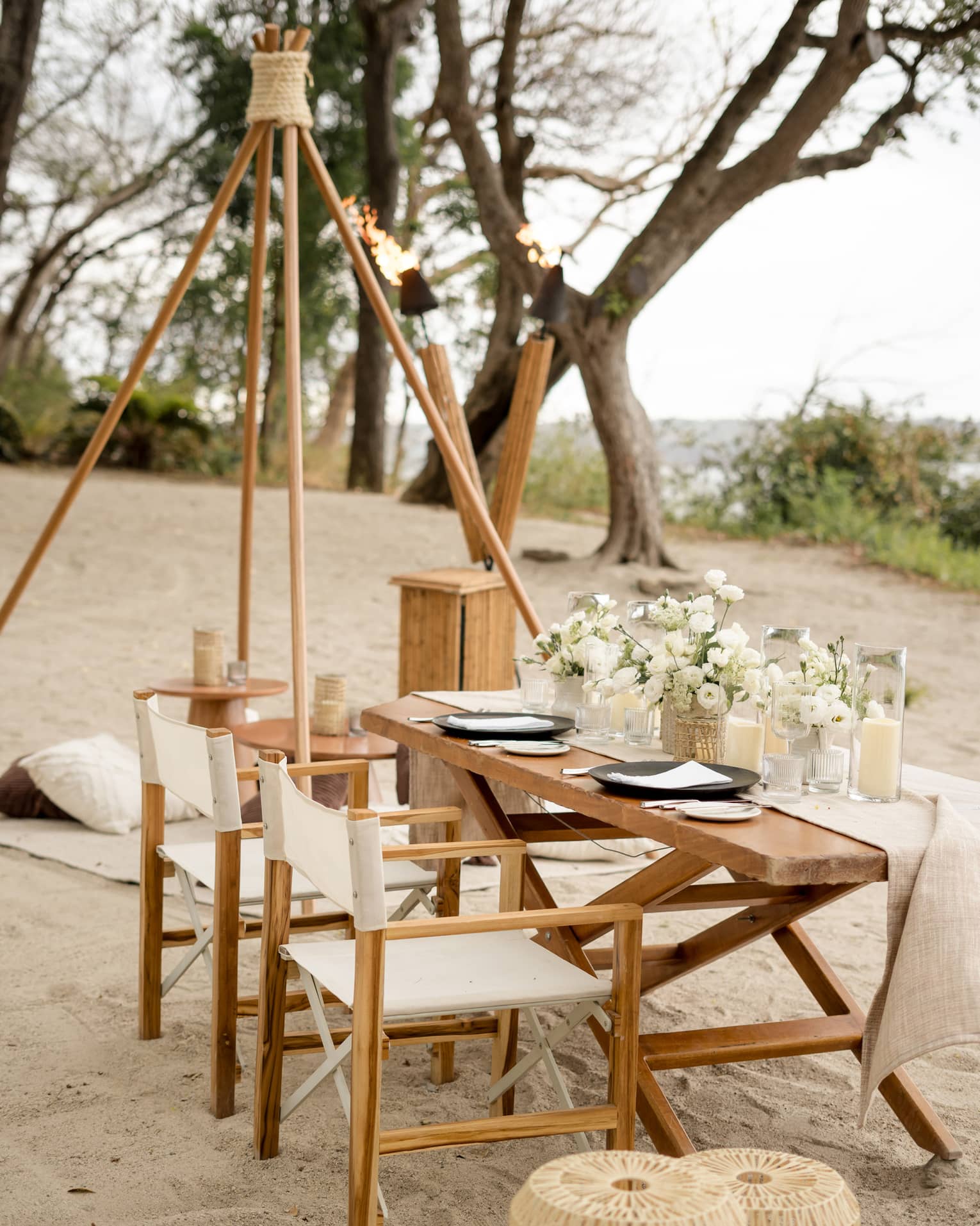 Wooden dining table and chairs set up on a beach, decorated with white floral centrepieces and candles