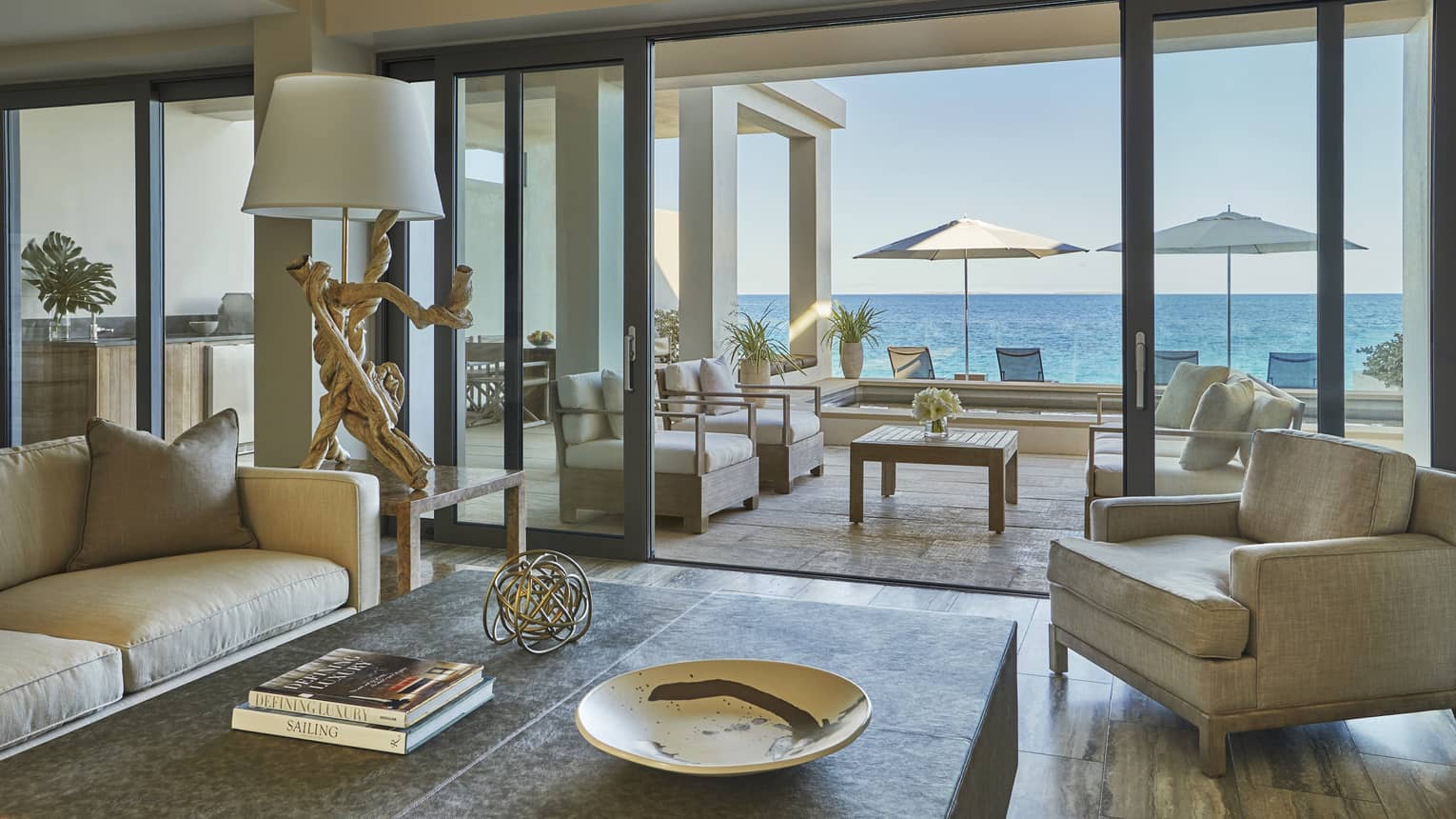 Elegant living room with neutral-toned seating, a coffee table and sliding glass doors opening to a terrace with ocean views and shaded loungers