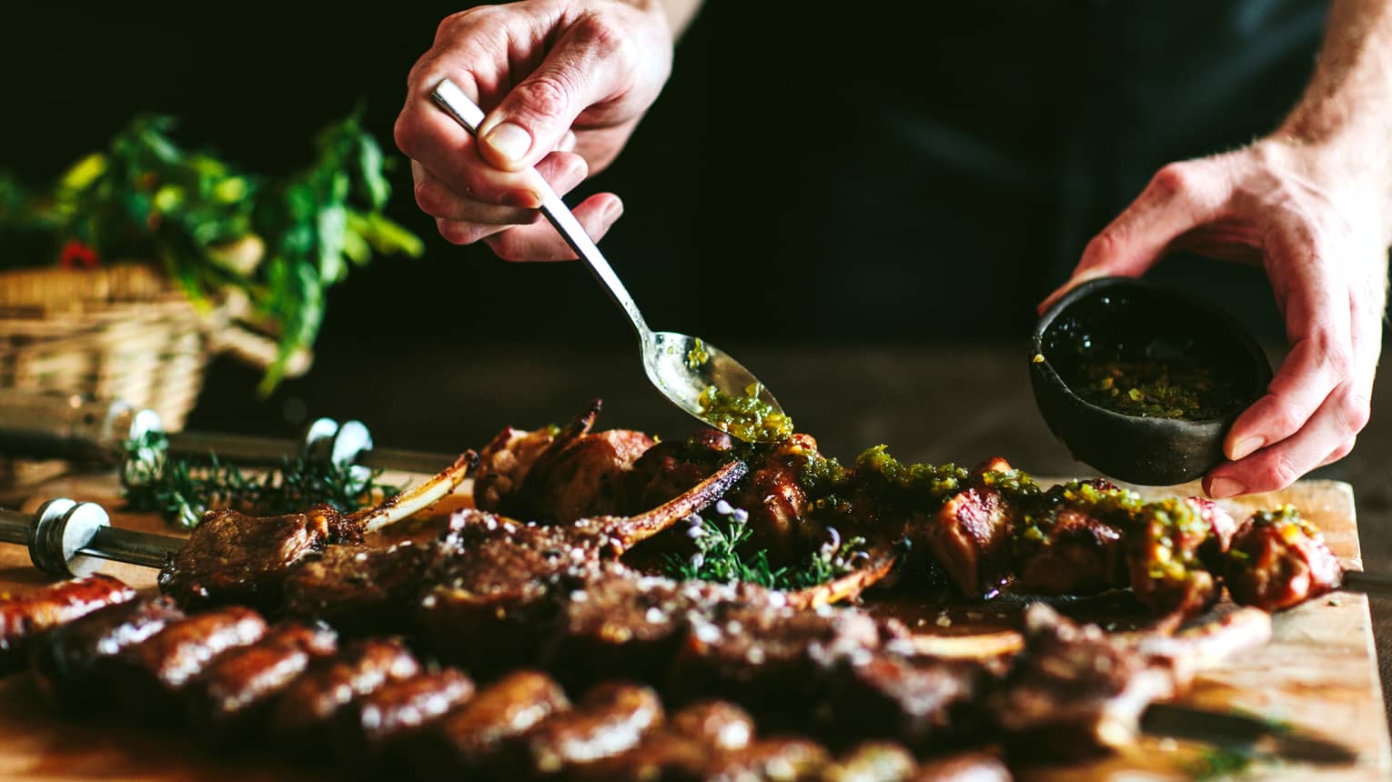 Man spoons sauce in small bowl over skewer of grilled meat 