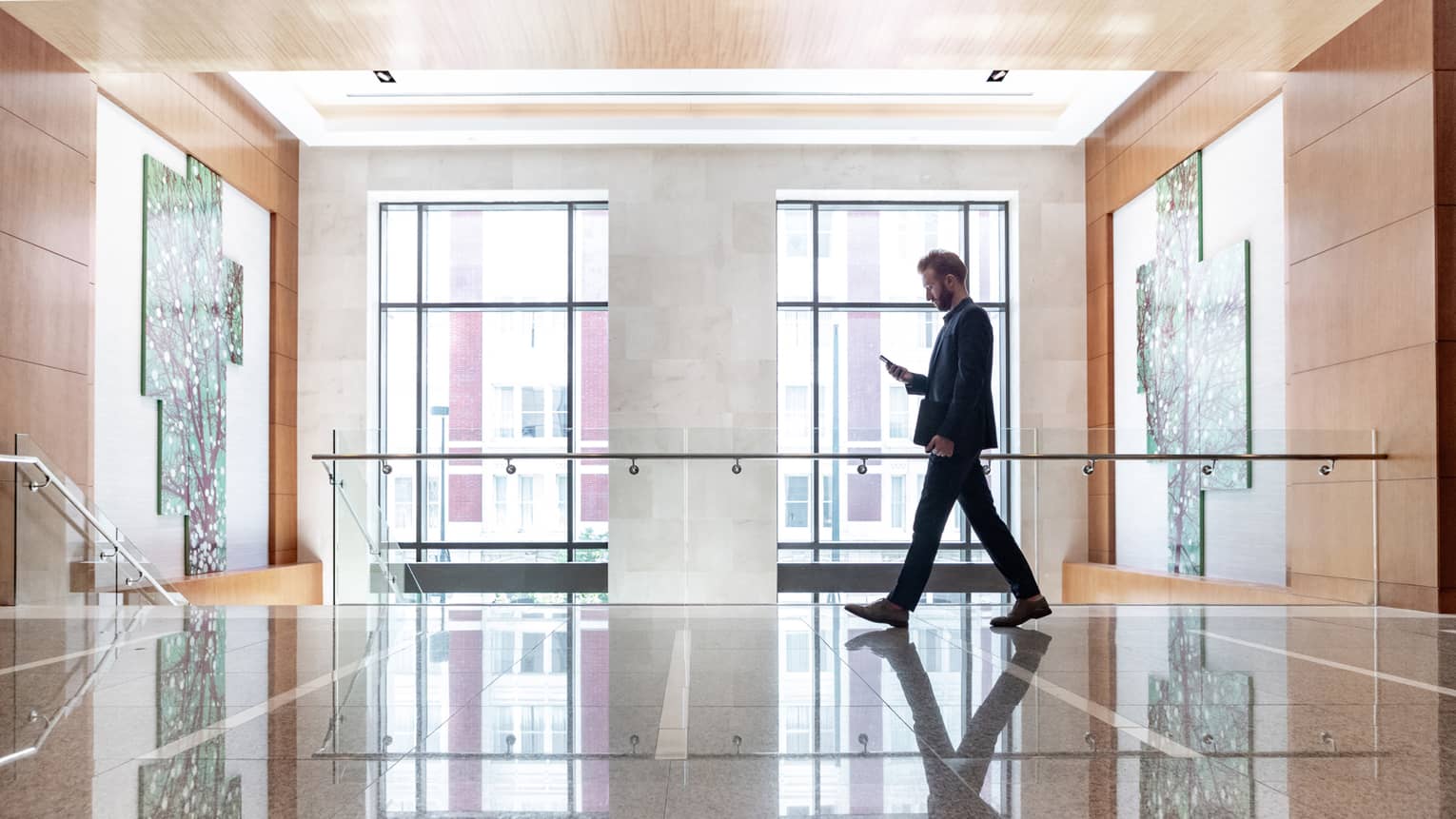 A man looking at his phone while he walks across a lobby area with large windows behind him