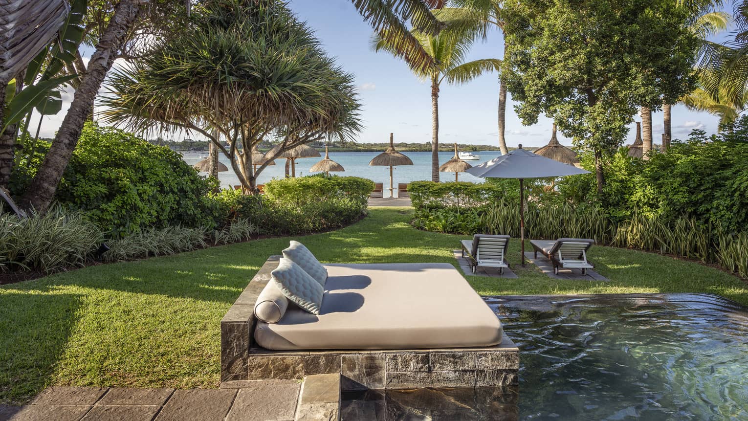 Tropical lawn with pool and day bed, two lounge chairs and umbrellas facing the beach