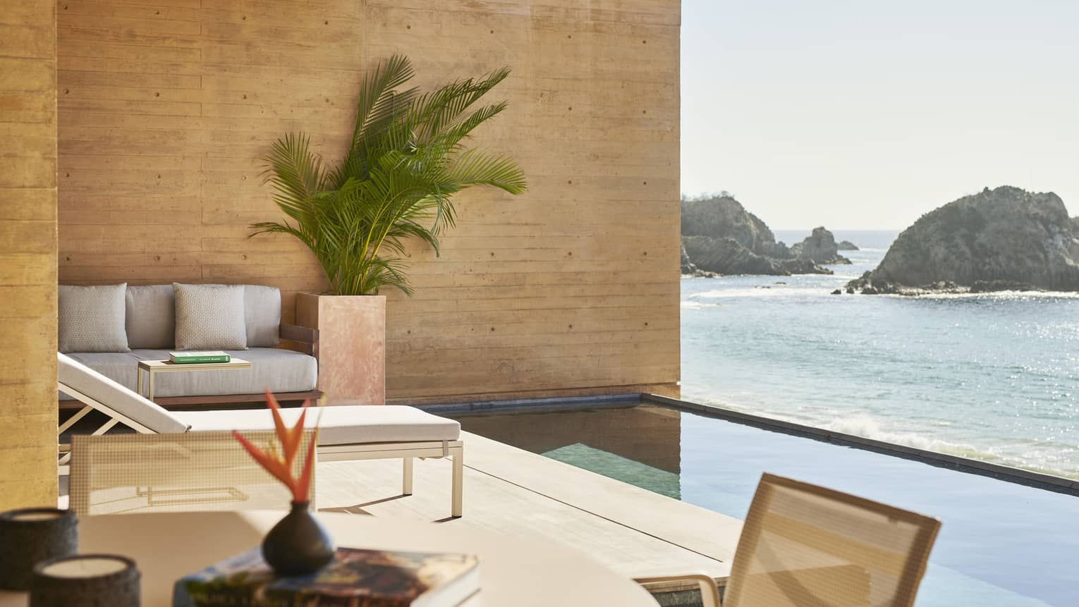 Outdoor seating area with modern furniture and tropical plants, overlooking an infinity pool and rocky coastline under clear skies. The space features natural wood walls.