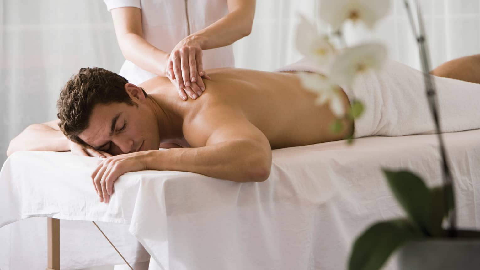Man lays on white table with eyes closed as masseuse massages his bare shoulders
