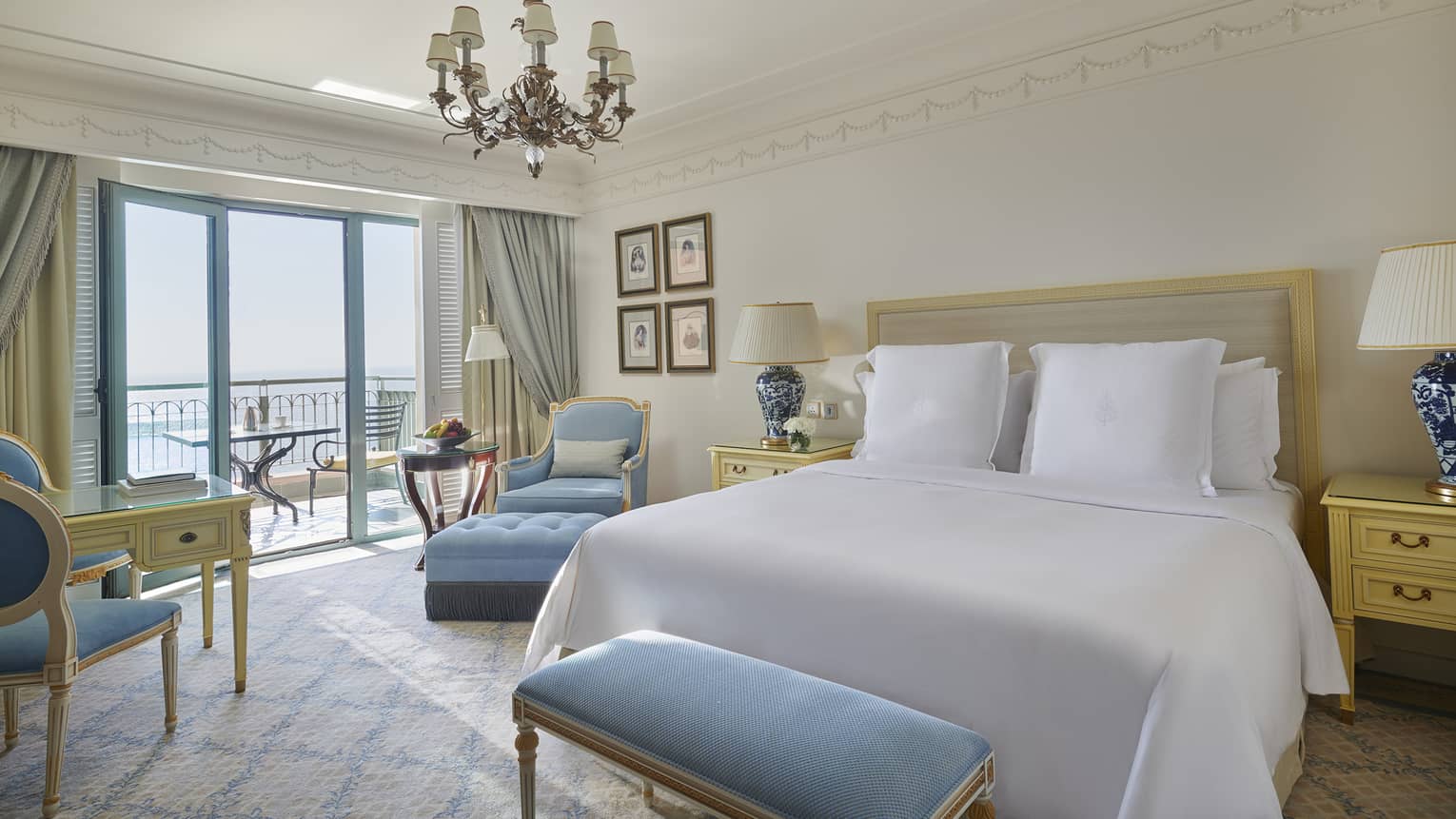 Bedroom with white king bed, chandelier, light blue bench and arm chair, and a balcony overlooking the sea