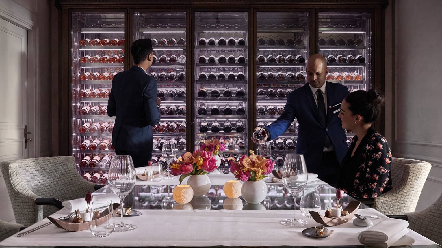 Man scans glass cellar as waitstaff pours wine in glass for woman at table