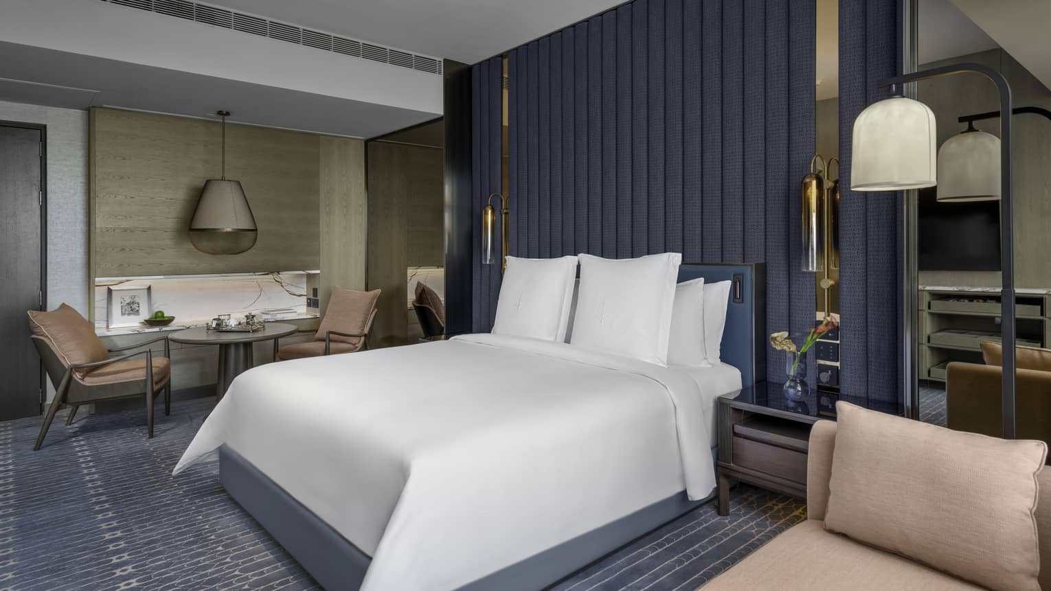 Renovated Deluxe City-View Room with bed and white linens, blue carpet and area rug, salmon chaise lounge, table for two with salmon armchairs