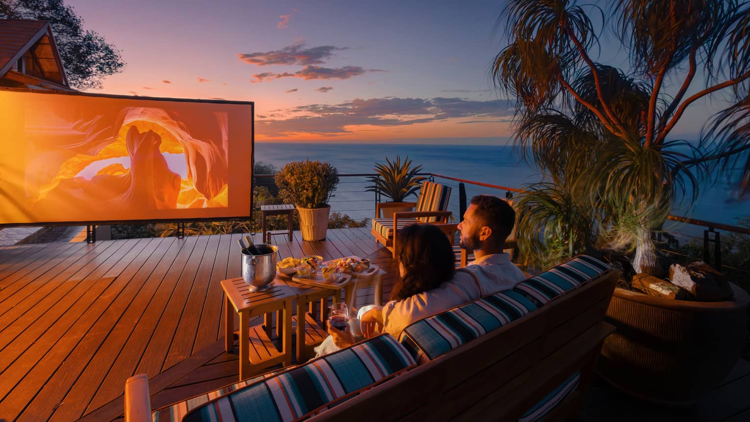 Two people sit on an outdoor sofa with a small table of food infront of them as they watch a movie on a large outdoor screen set up on a private terrace