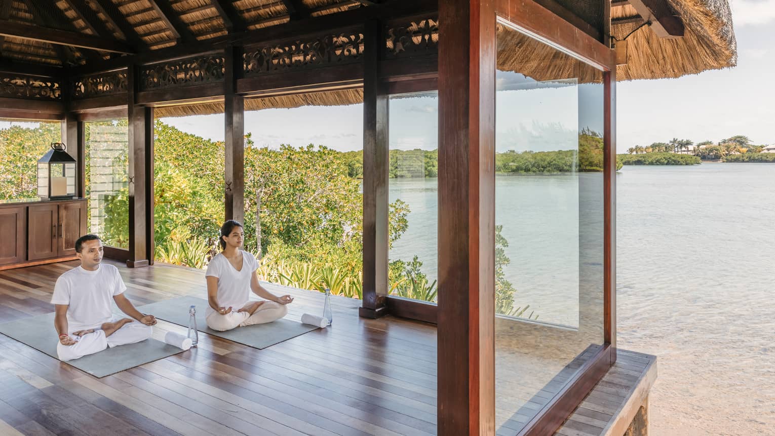 Two people wearing white seated in yoga poses in open-air spa yoga pavilion by beach