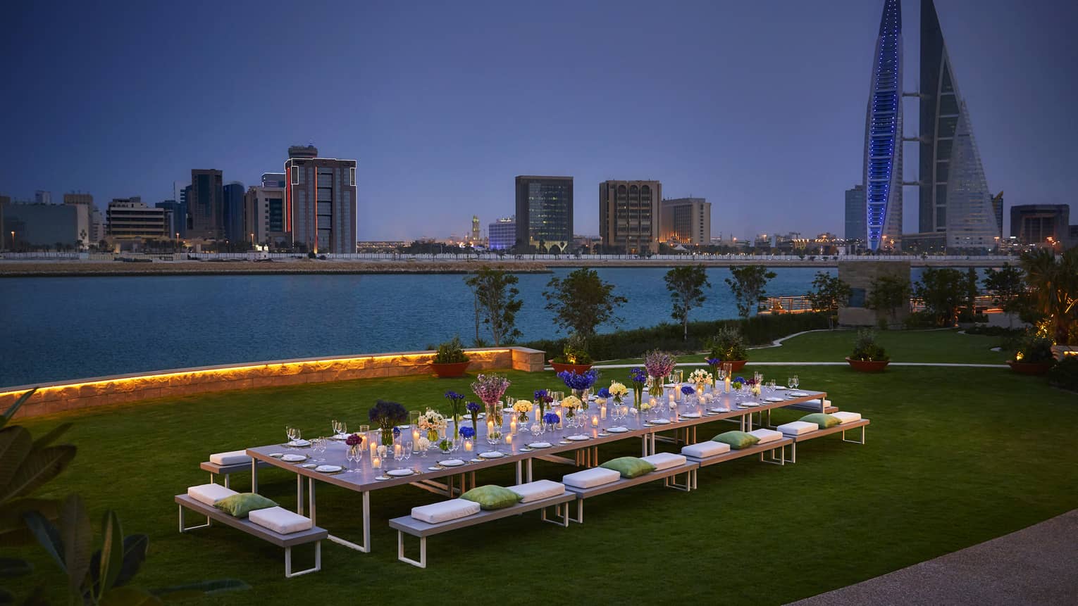 Long, candle-lit dining table on event lawn by water at night