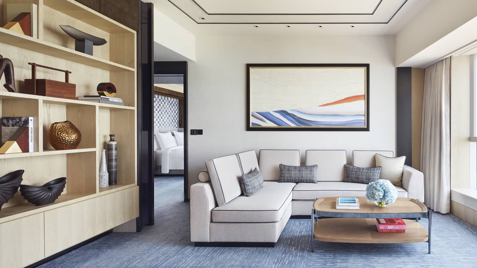 Modern hotel living area with beige sectional sofa, abstract wall art, open shelving with decor and coffee table with books and blue flowers.