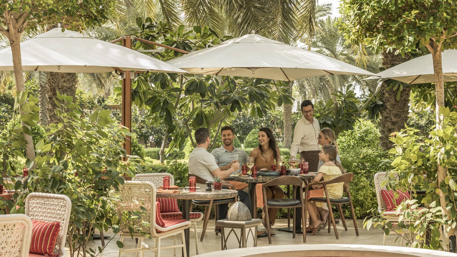 A group of people laugh at a wrought iron table in a lush green garden.