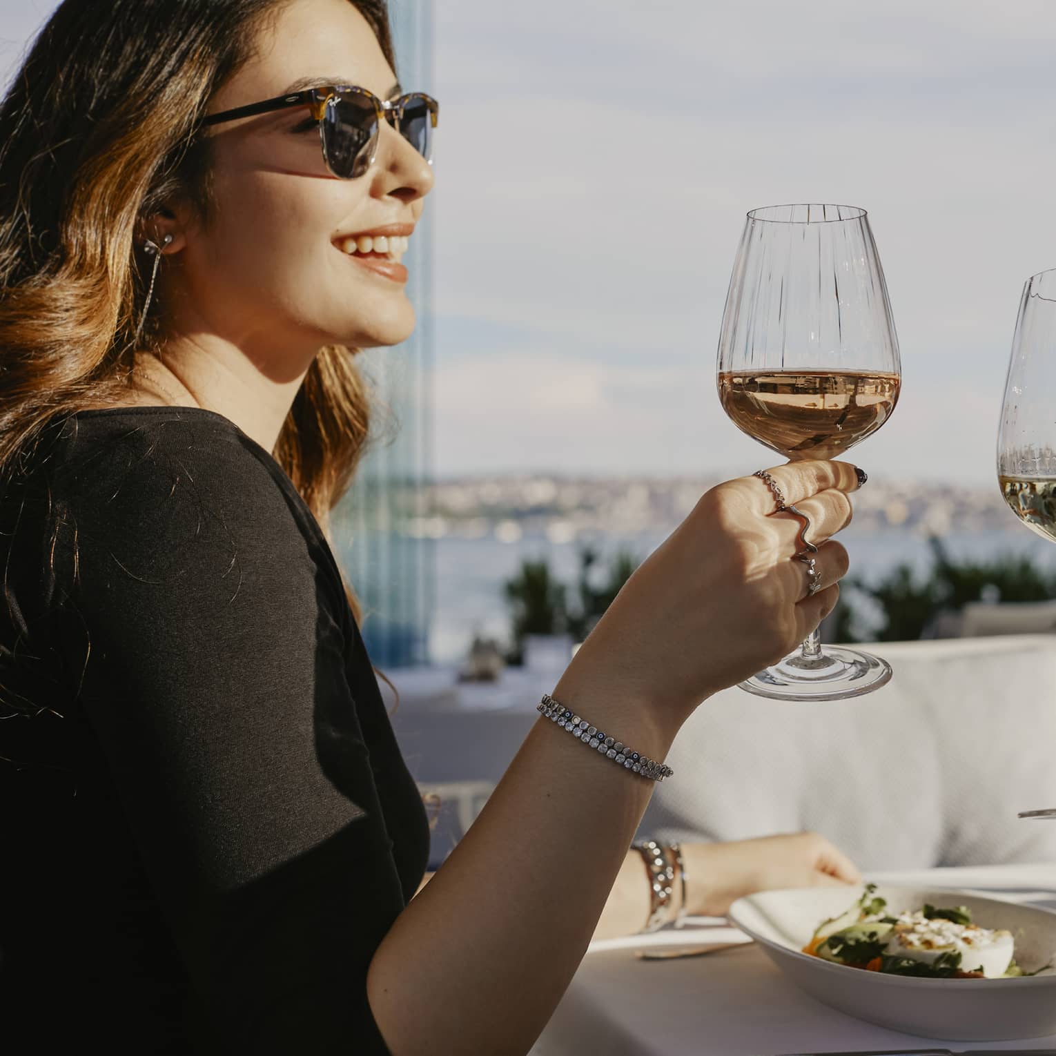 Person wearing sunglasses, smiling while toasting with a glass of wine with another person