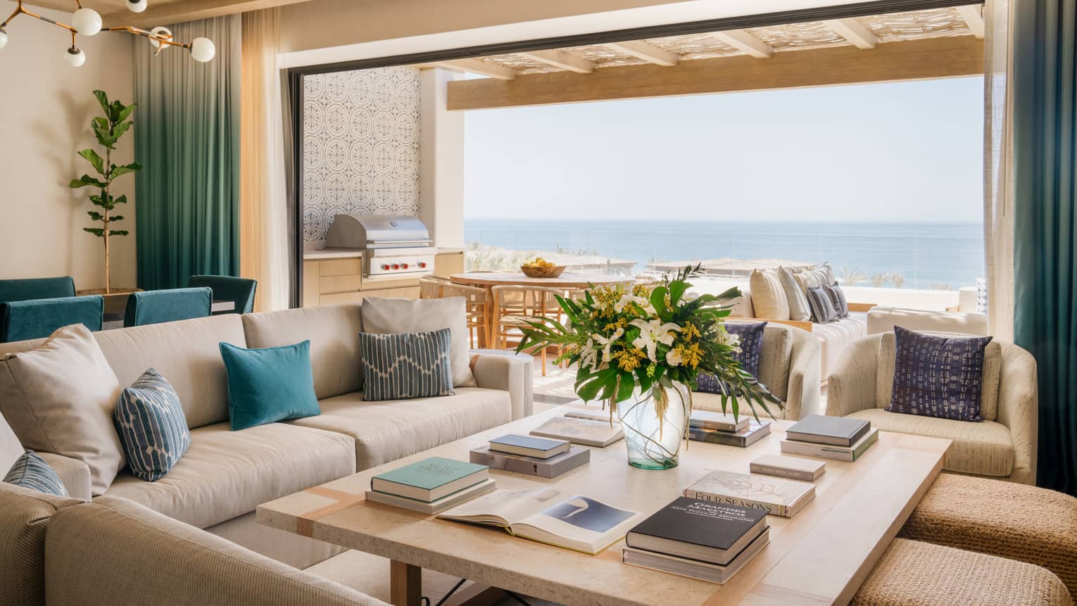 Luxury villa living room with sofa, chairs and ottomans around a large square coffee table, with a wall open to ocean-view pool terrace, at Four Seasons Resort Cabo San Lucas