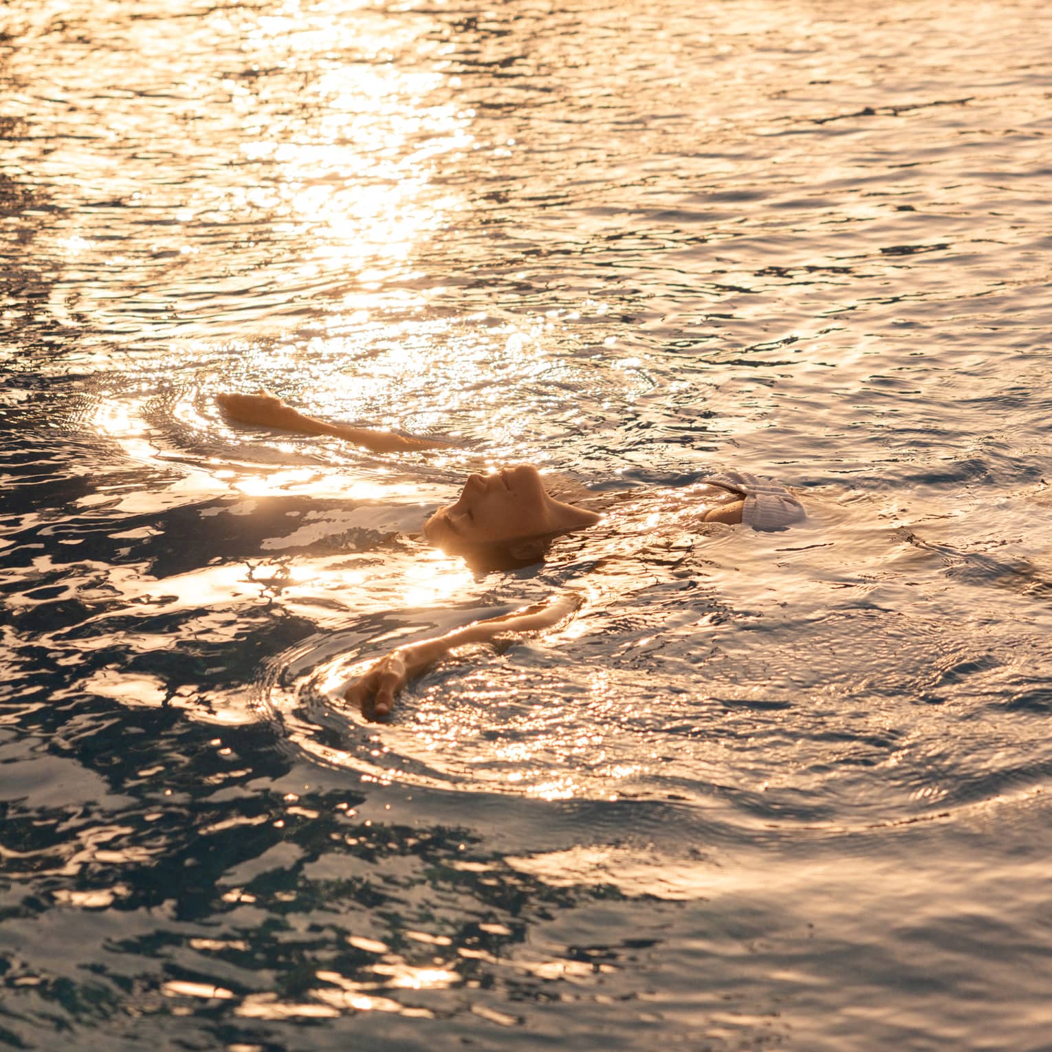 A person floating on their back in the ocean, with sunlight rippling across the water.