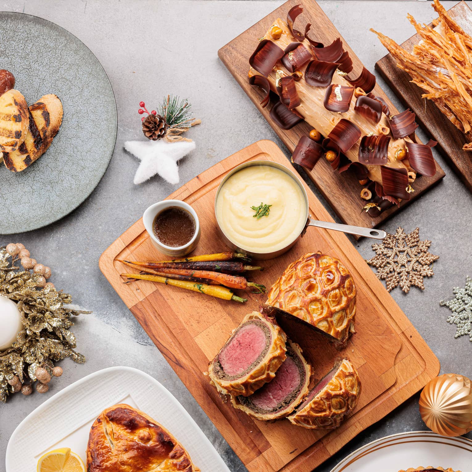 Overhead view of an assortment of dishes including a sliced beef Wellington, whole-roasted fish, yulie log and other desserts all on a light-grey table