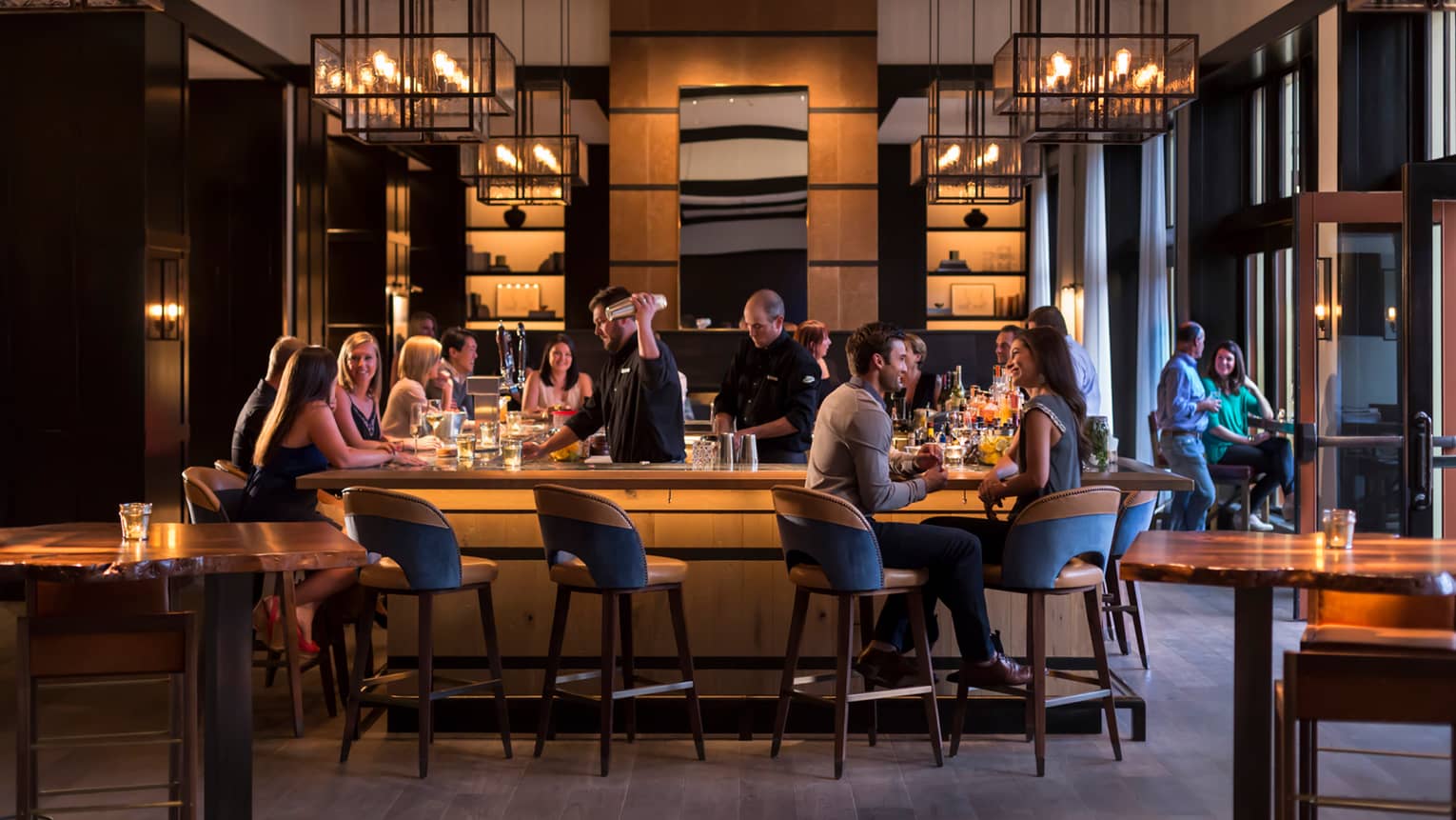 Guests socialize around wood bar with mid-century style stools and chandeliers as bartender makes martini