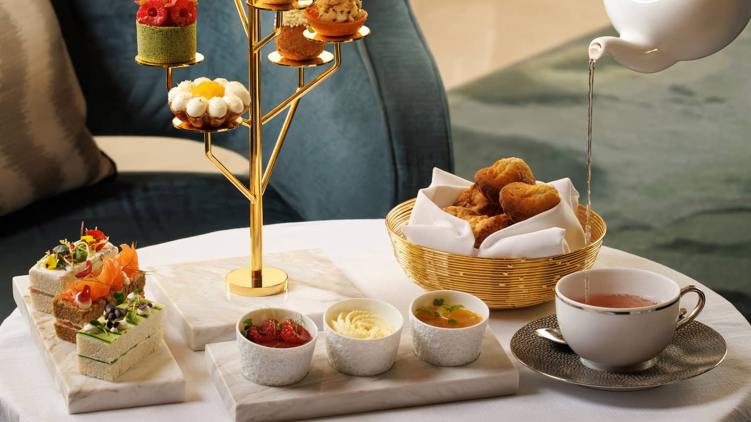 Golden tree-shaped stand holds a small pastry on each branch, set next to a marble plate of finger sandwiches, basket of bread, three small white bowls and a cup of tea