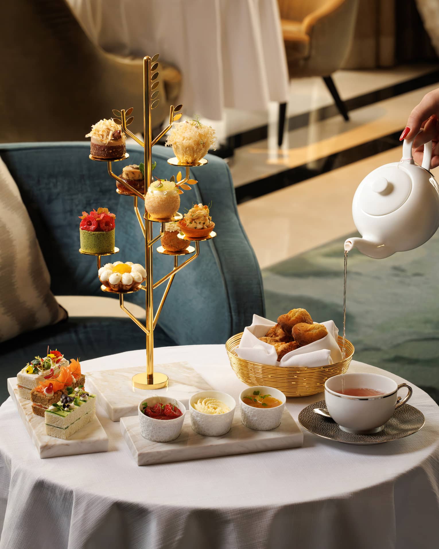Golden tree-shaped stand holds a small pastry on each branch, set next to a marble plate of finger sandwiches, basket of bread, three small white bowls and a cup of tea