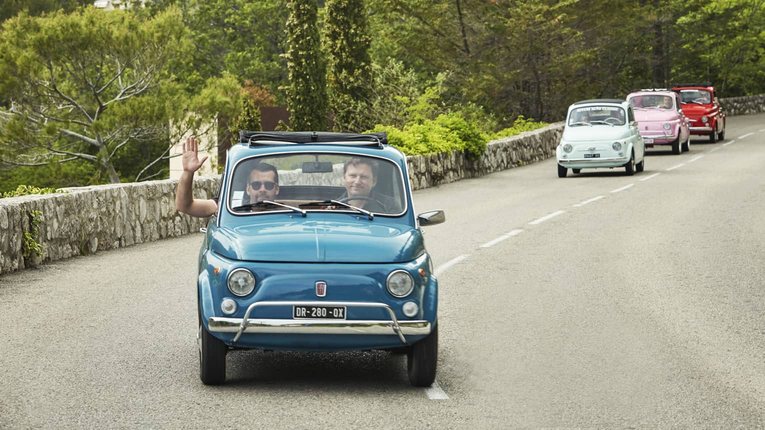Tourists drive colourful Fiat 500s down a road lined with a stone wall and greenery. The passenger of the first car waves.