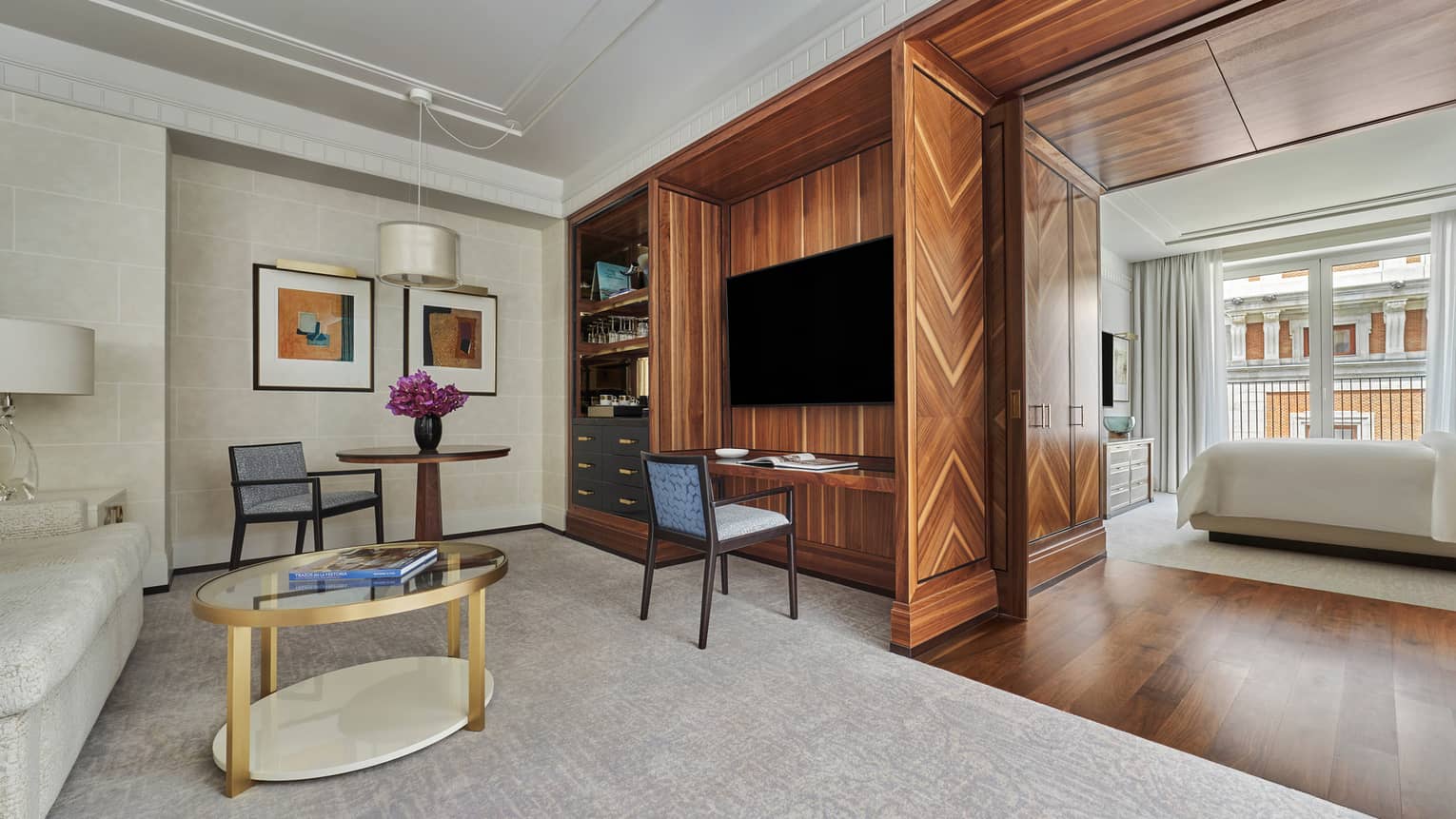 Luxury hotel suite living area featuring a wood-panelled TV wall, sofa, round tables and modern chairs, leading to a bedroom with floor-to-ceiling windows
