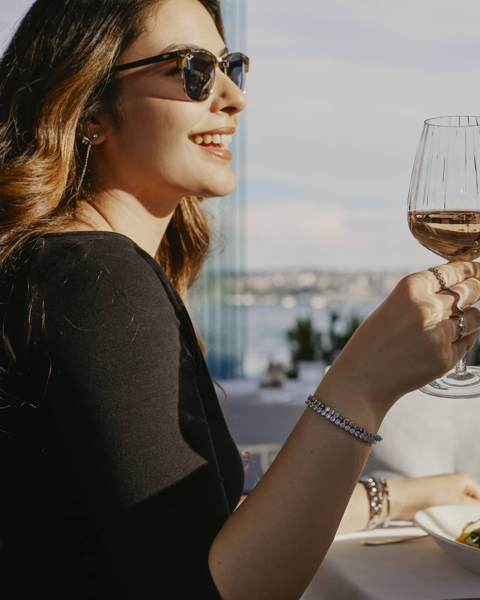 Person wearing sunglasses, smiling while toasting with a glass of wine with another person