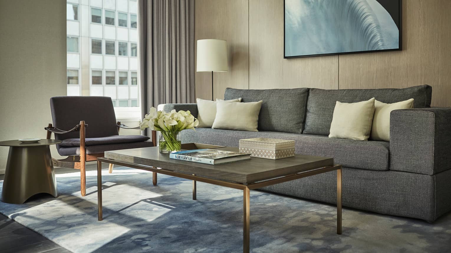 Hudson Suite long grey sofa, table with white flowers, books, armchair by window, building view