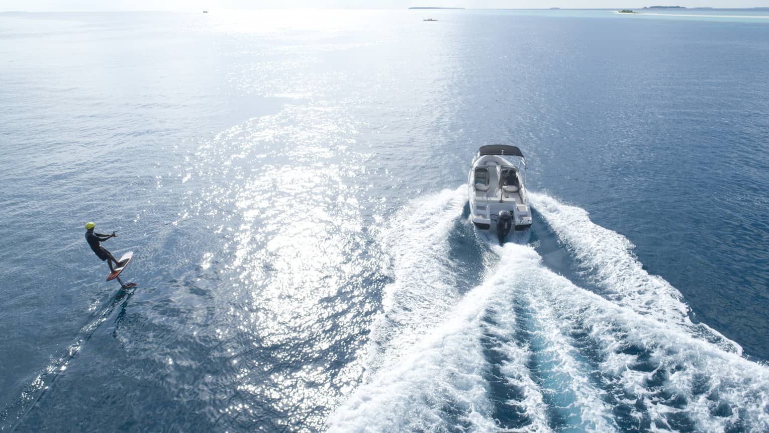 Aerial view of a motorboat towing a wakeboarder, churning up white waves as it speeds through calm water. White sky overhead.