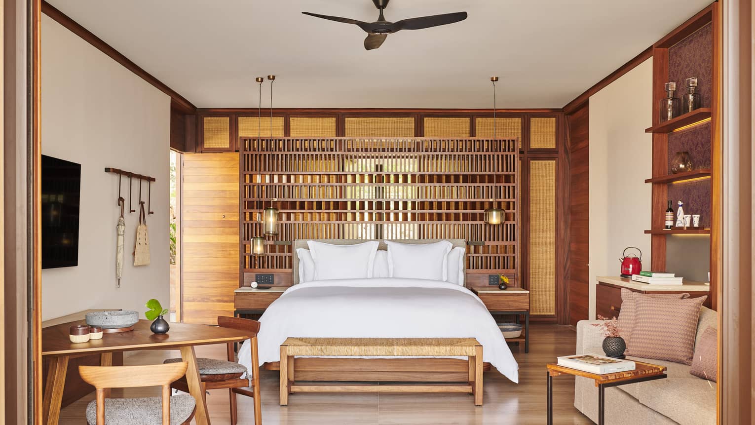 A stylish bedroom with a large bed, wooden lattice headboard and wicker bench. The room includes a small dining table, cozy seating area and warm wooden accents throughout.