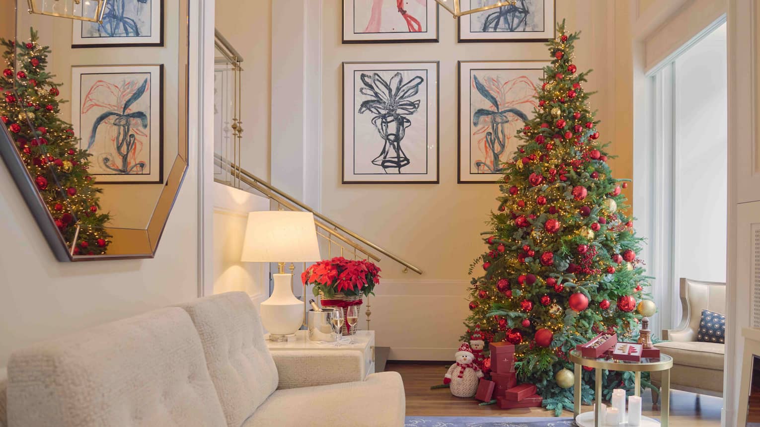 A spacious guest room with pale neutral decor, an off-white couch, an end table with a table lamp and vase of flowers, an upholstered chair, a large mirror and wall art. A decorated Christmas tree stands in a corner.