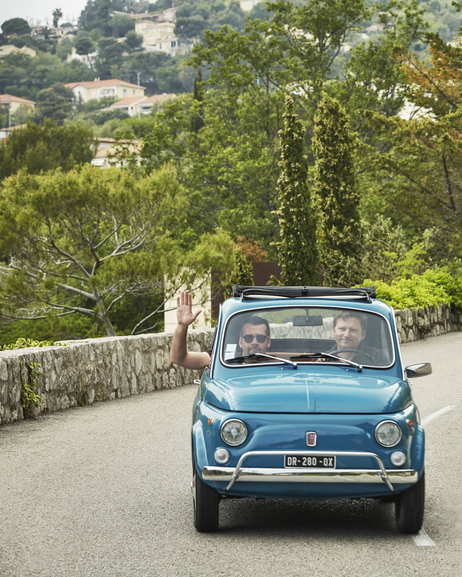 Tourists drive colourful Fiat 500s down a road lined with a stone wall and greenery. The passenger of the first car waves.