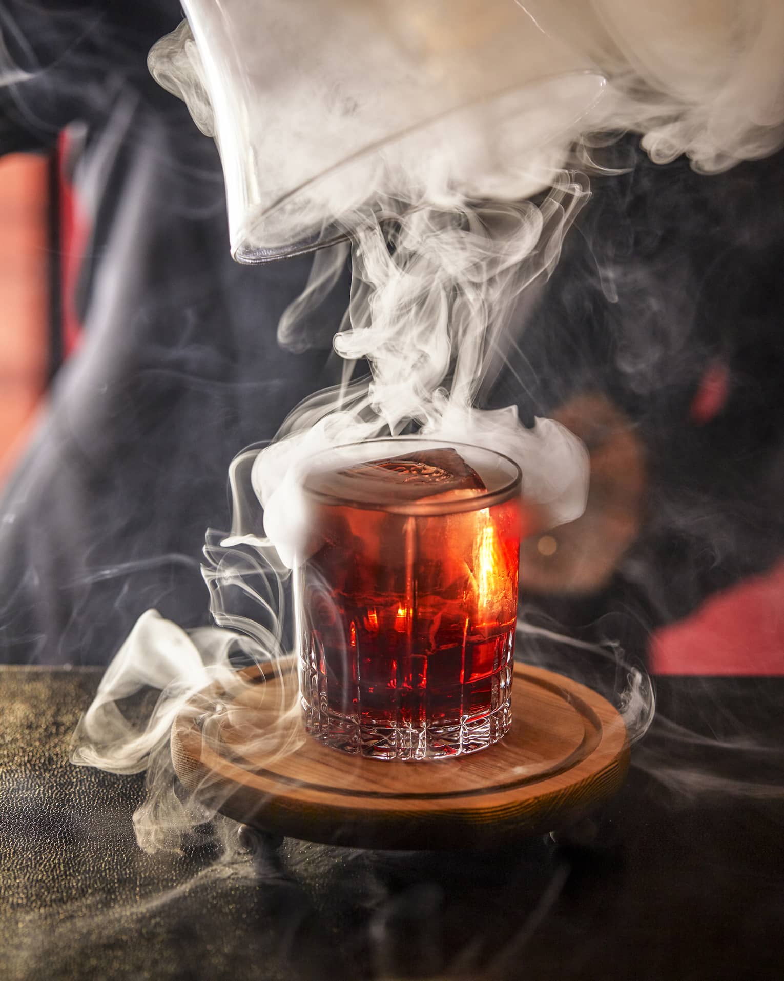 Bartender swirls smoke on top of a shot glass of amber-coloured liquor 
