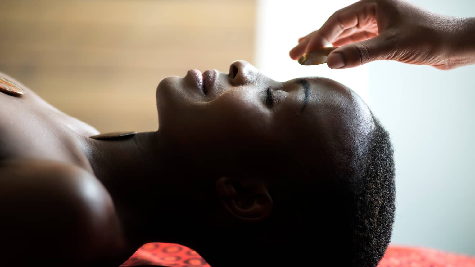 Hand placing stone on woman's forehead as she smiles, face-up on spa table