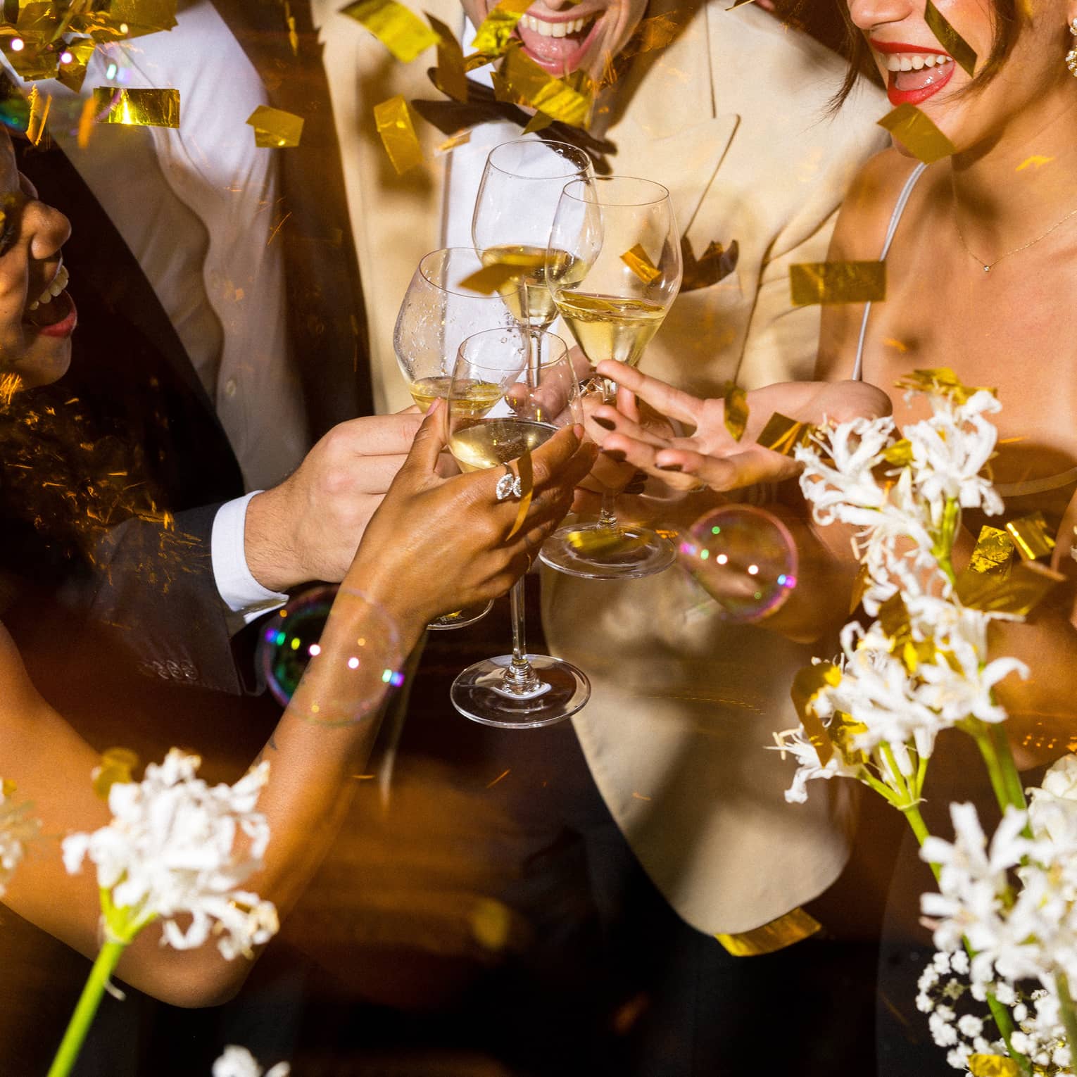 Four people toast glasses of champagne surrounded by gold confetti and white flowers