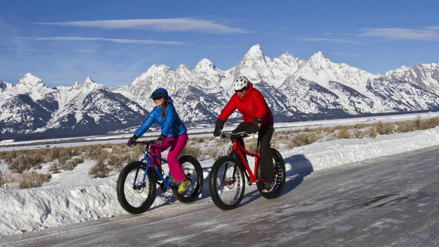 A man and woman bike down a path, snow-capped mountains and blue sky in the background