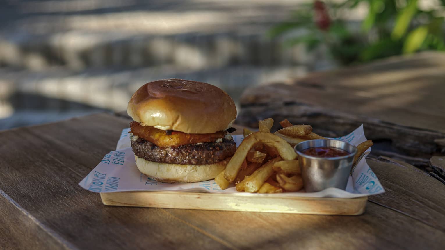 A small rectangular wooden platter holds a hamburger in a bun, French fries and a small steel container of ketchup on a wooden picnic table