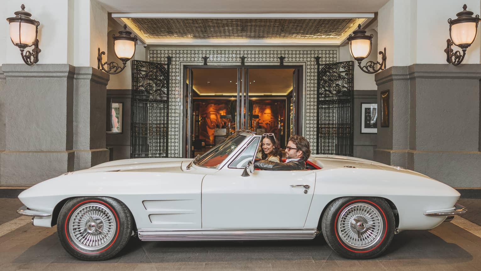 A couple sitting in a vintage white convertible parked outside a luxury hotel entrance, surrounded by ornate lighting and elegant decor.