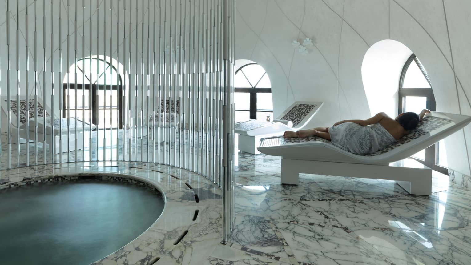 Back of woman wearing white towel, lying on white lounge chair next to small round pool in spa
