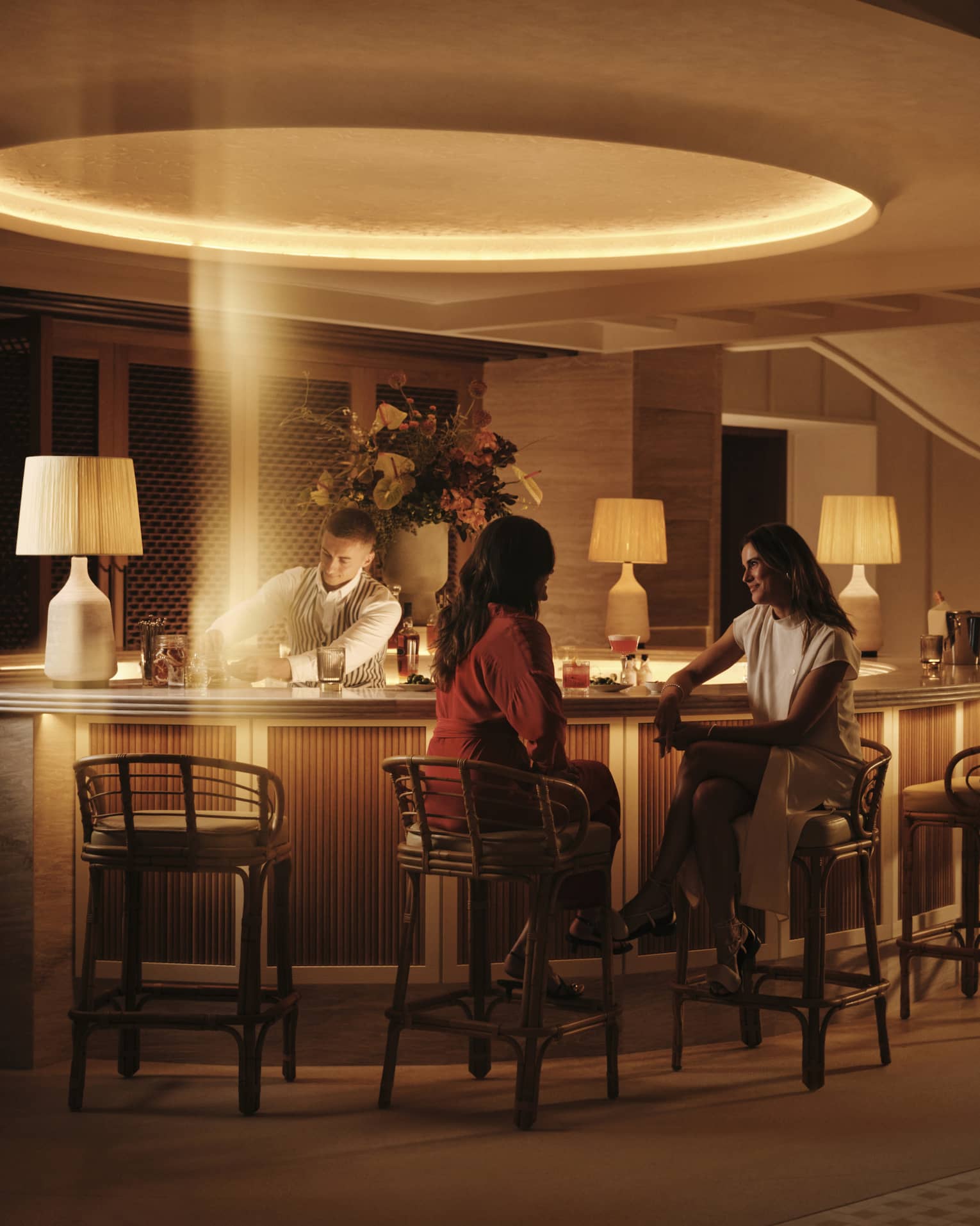Two smiling guests sit at a warmly lit circular bar at night as a bartender prepares cocktails in a stream of golden light.