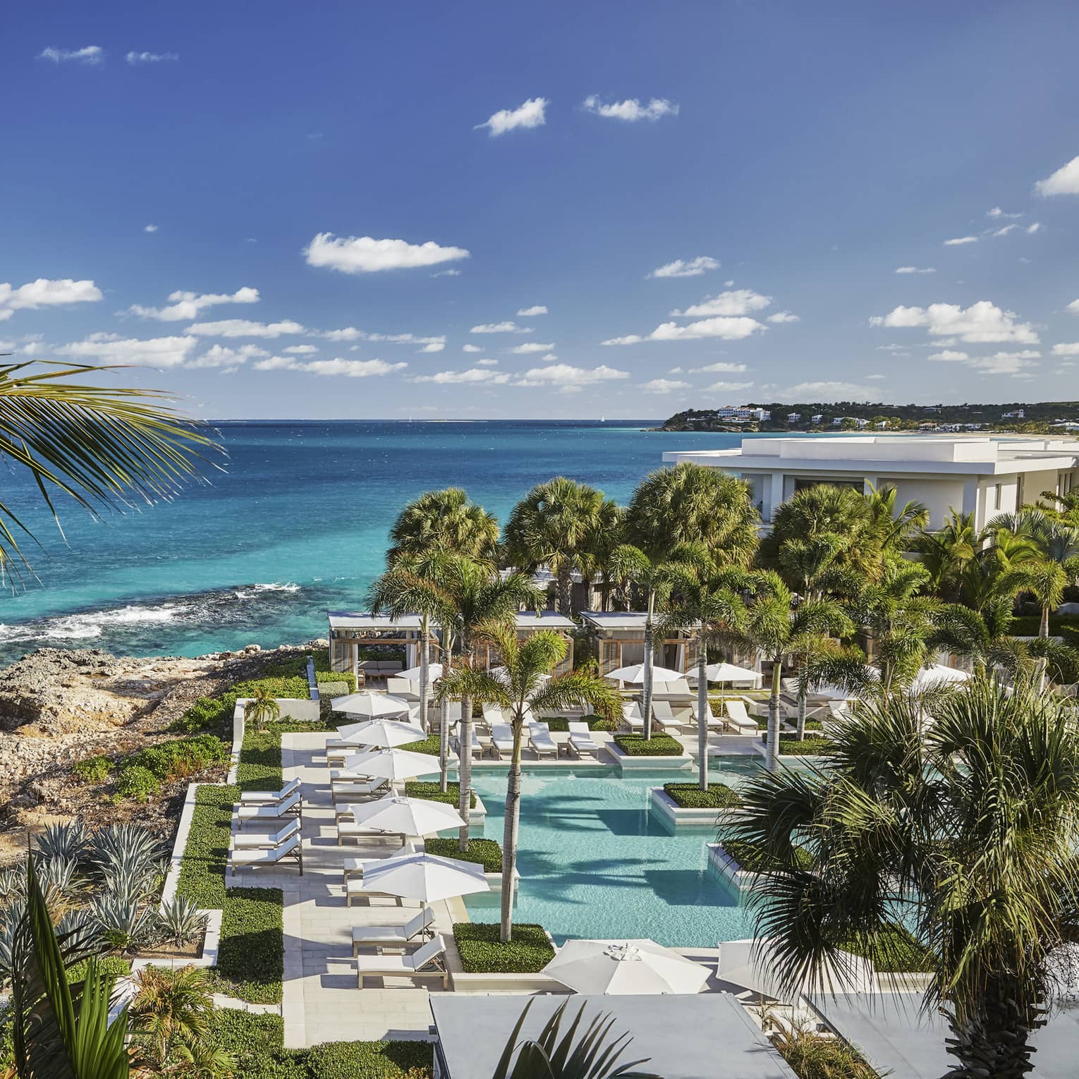Looking down over Anguilla outdoor pool, patio, large rocks along blue-green ocean waterfront, blue sky
