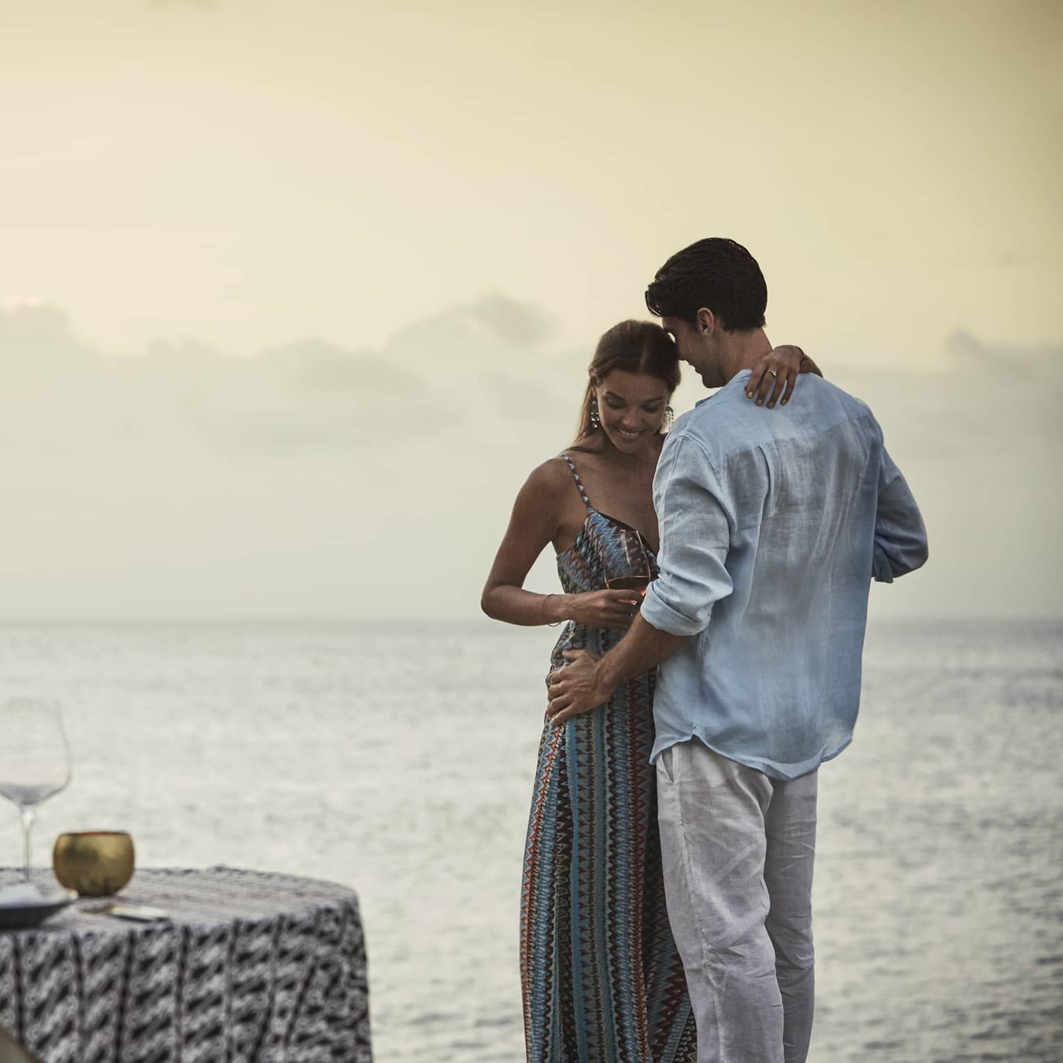 Couple embraces beside private dining table overlooking ocean at sunset