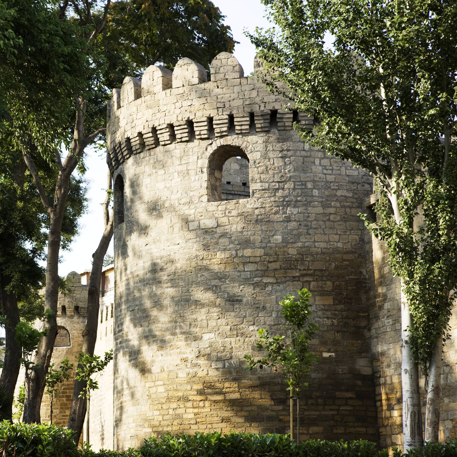 A cylindrical structure marked by loophole openings and grey stone exterior: part of a crenelated tower surrounded by trees. 