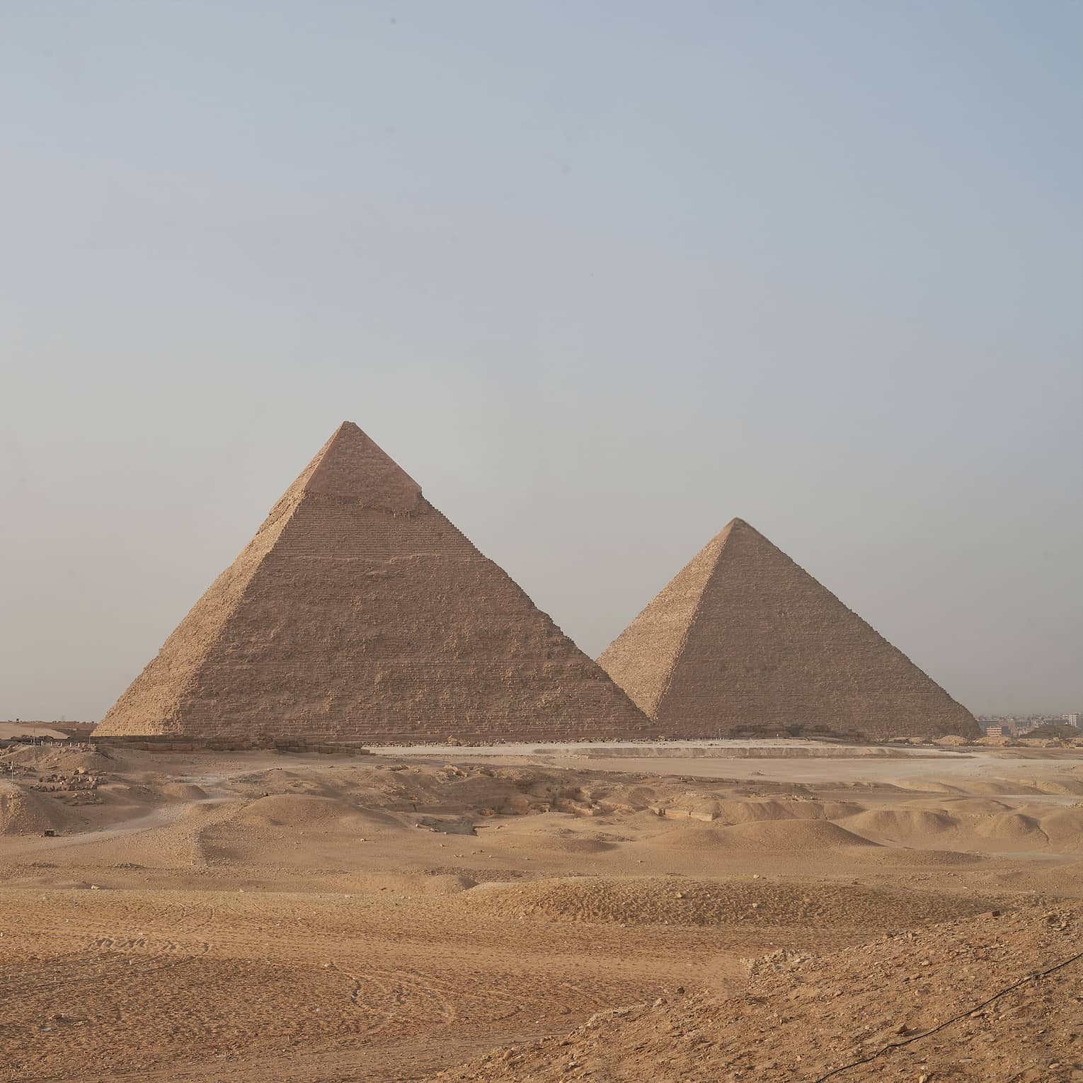 In the arid desert, a line of three towering pyramids dwarf two smaller pyramids before them, under a hazy blue sky. 