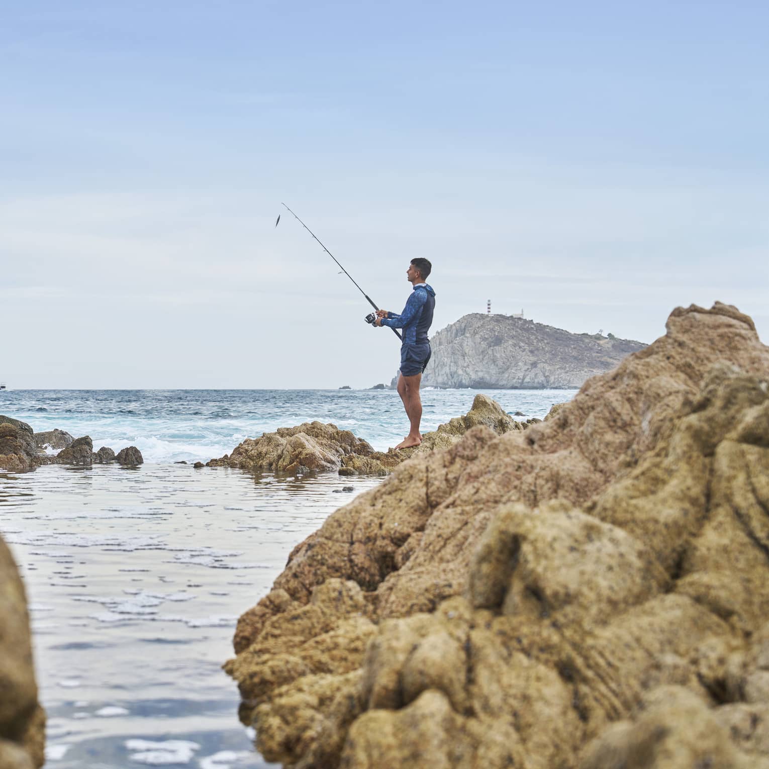 A fisher holding a pole stands barefoot atop a rocky outcrop and looks out toward an expanse of blue water under a clear sky.