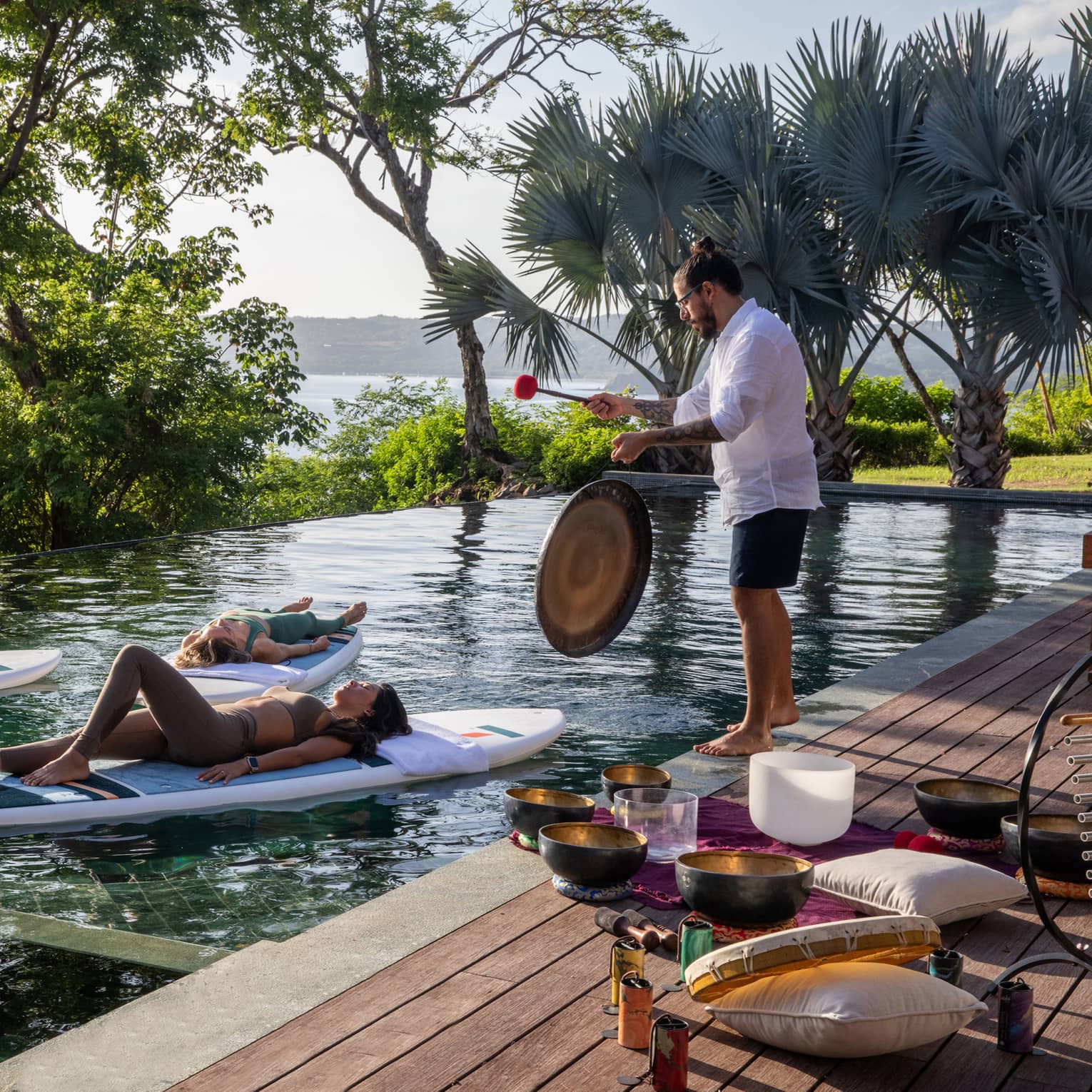 Three people float on stand-up paddleboards in a private pool while someone plays a gong on the deck