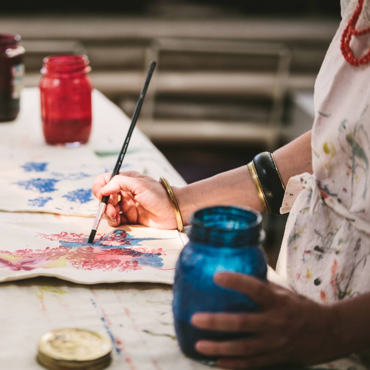 An artist using mason jars filled with red, blue, yellow and black paint to paint images of flowers on canvas tote bags.