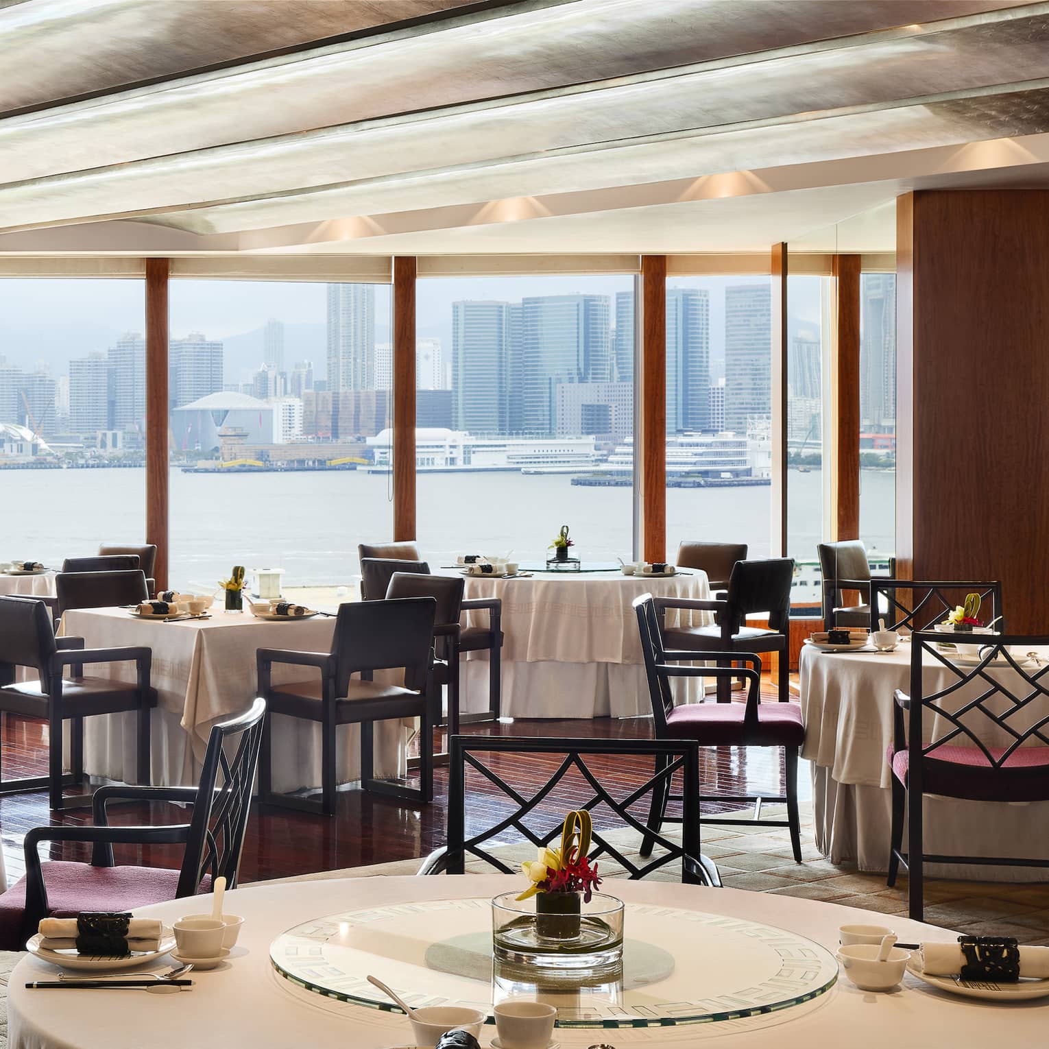 An elegant dining room with tables set with flowers and place settings, overlooking the city from across a body of water.