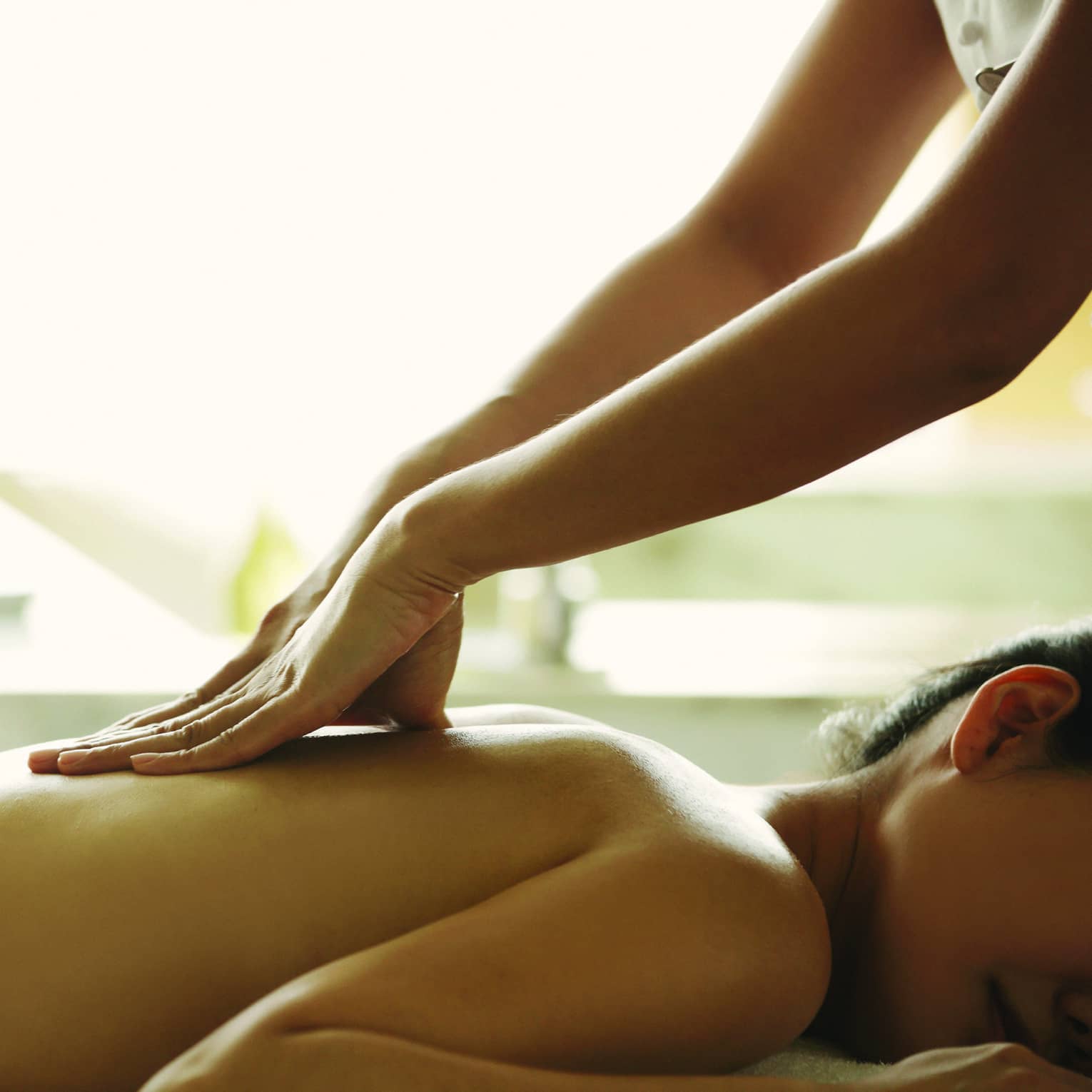 Masseuse reaches over, massages woman's bare back as she lies on spa table
