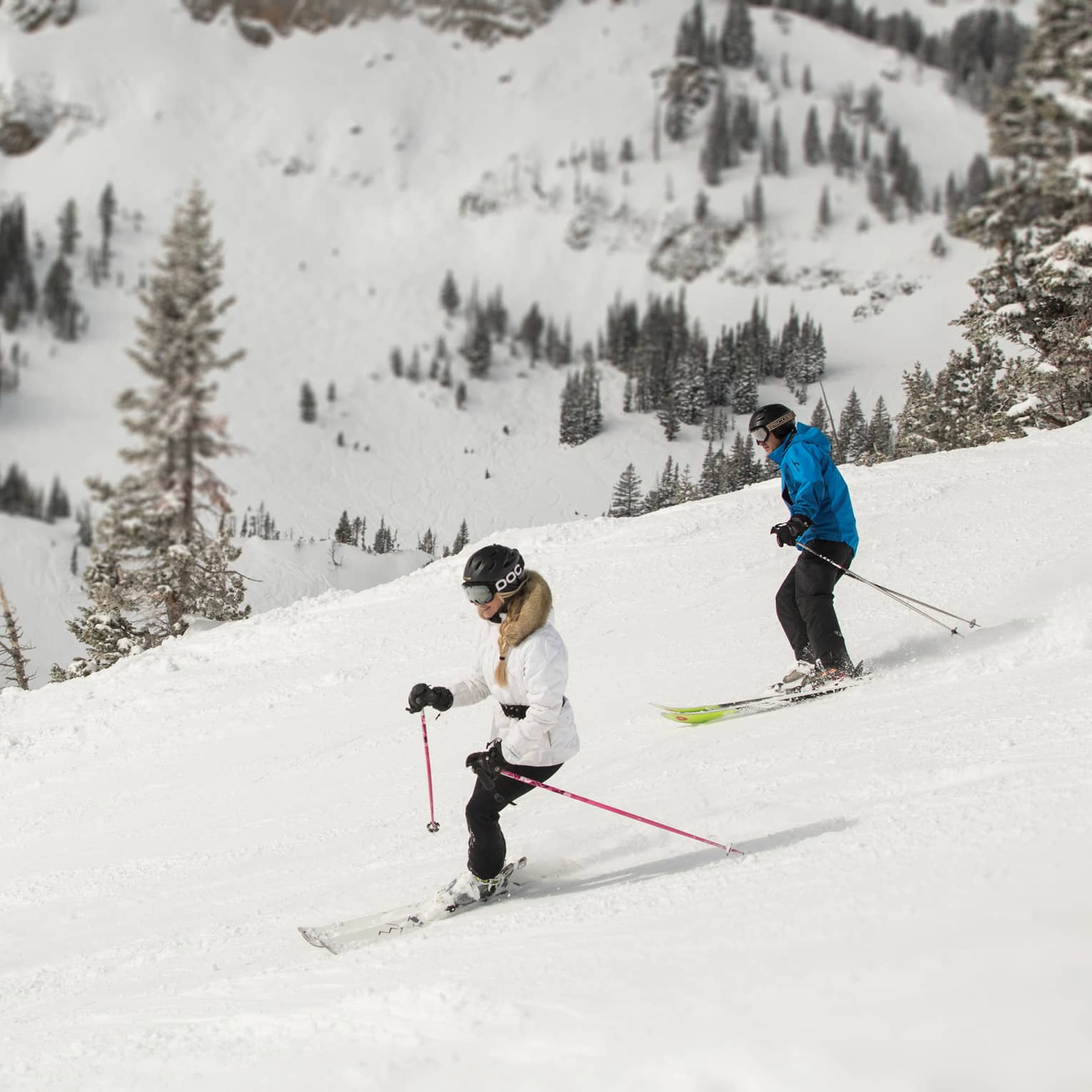 Three skiers descending a snowy mountain slope, surrounded by snow-covered trees and rugged terrain